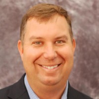 Stanislaus Tuholski, a brown-haired person wearing a suit, poses against a gray background for a headshot.