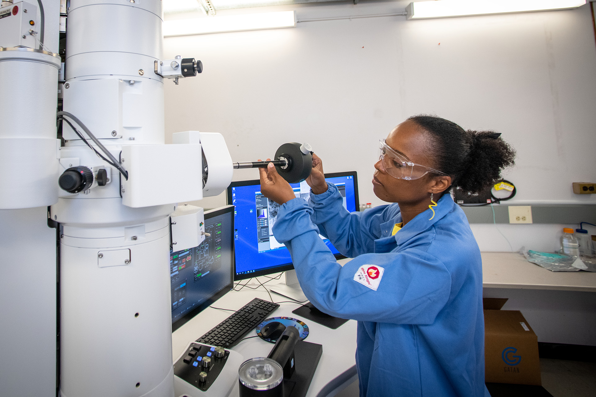 Dark-haired scientist in a blue lab coat adjusts a large piece of machinery with a tool.