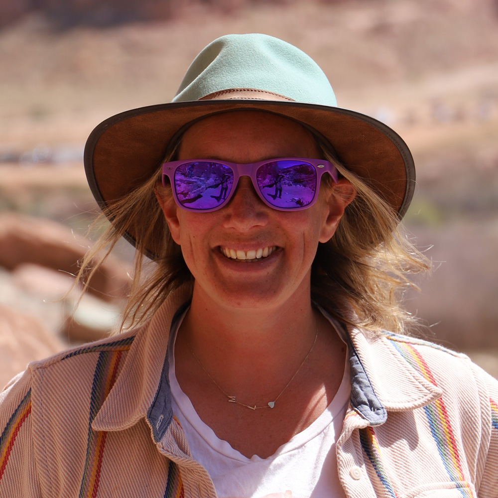 Person with medium length blonde hair wearing a hat, sunglasses, and a collared tan jacket over a white shirt, photographed outdoors.