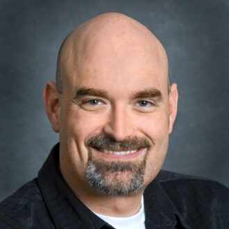 Blake Simmons, a person with a gray beard wearing a black jacket over a white shirt, photographed against a gray backdrop.