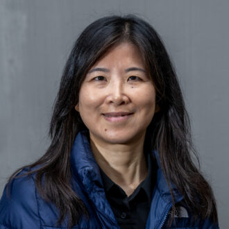 A person with long dark hair wearing a blue puffer jacket over a black top. Photographed against a gray background.
