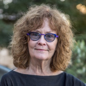 A cropped portrait of Carolyn Larabell, a person with curly shoulder-length brown hair wearing blue glasses and a dark top, smiling.