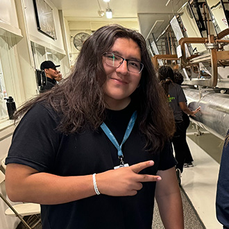 Person with long brown hair wearing glasses, a black shirt, and a blue lanyard, holding up a peace sign, smiling.