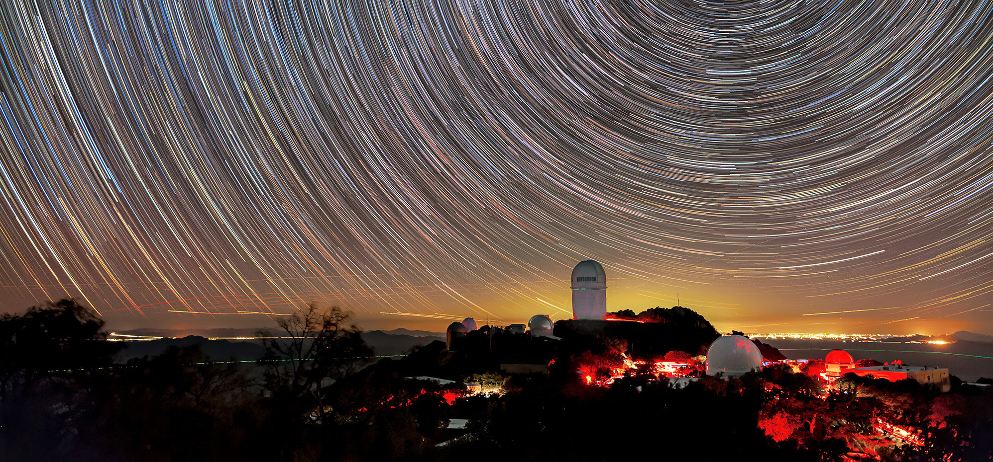 A long-exposure image captures circular star trails above telescope domes illuminated in red light on a dark mountain. A golden glow on the horizon marks distant city lights.
