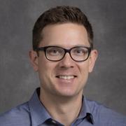 Stefan Minasian, a person with short brown hair wearing a blue collared shirt and black glasses, smiling in front of a gray backdrop.