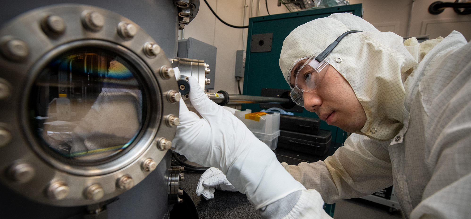 A scientist in a cleanroom suit, gloves, and safety glasses carefully examines a vacuum chamber, with their reflection visible in a round glass window.
