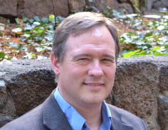 Dan McKinsey, a person with short light blonde hair wearing a navy blazer over a blue collared shirt, photographed against a stone wall with greenery.