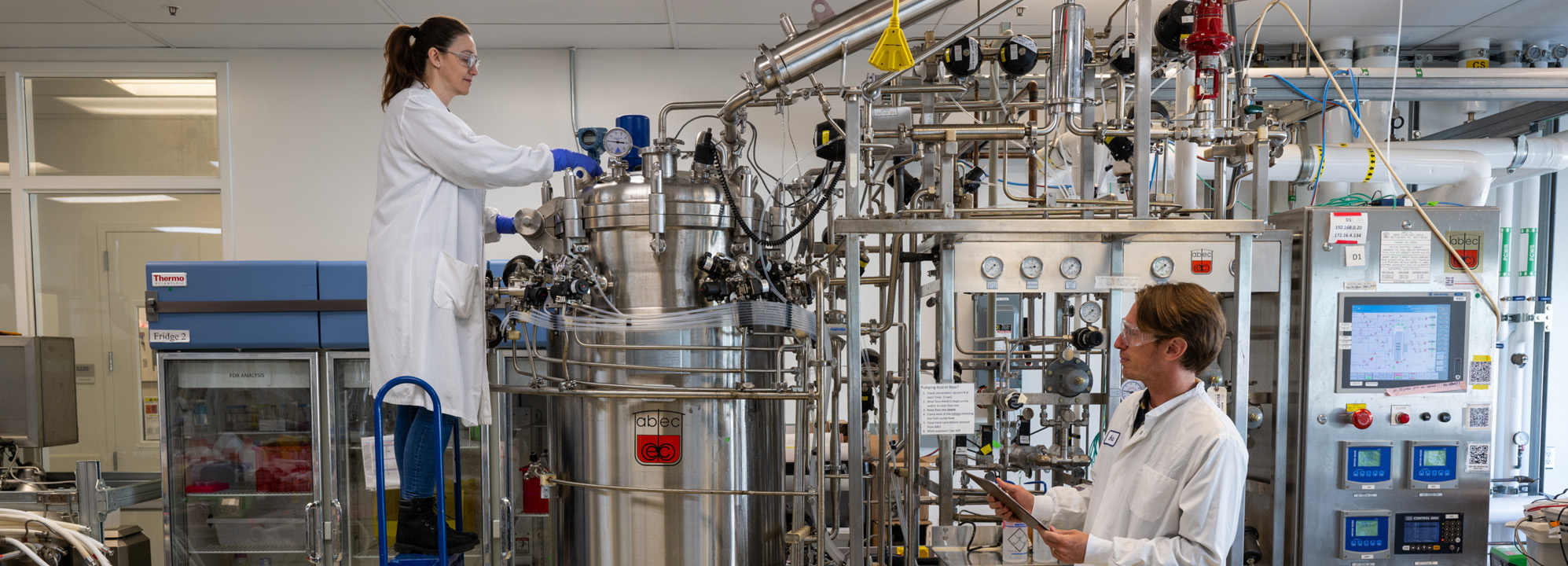 Two researchers wearing white lab coats and safety goggles working with large machinery in the Lab.
