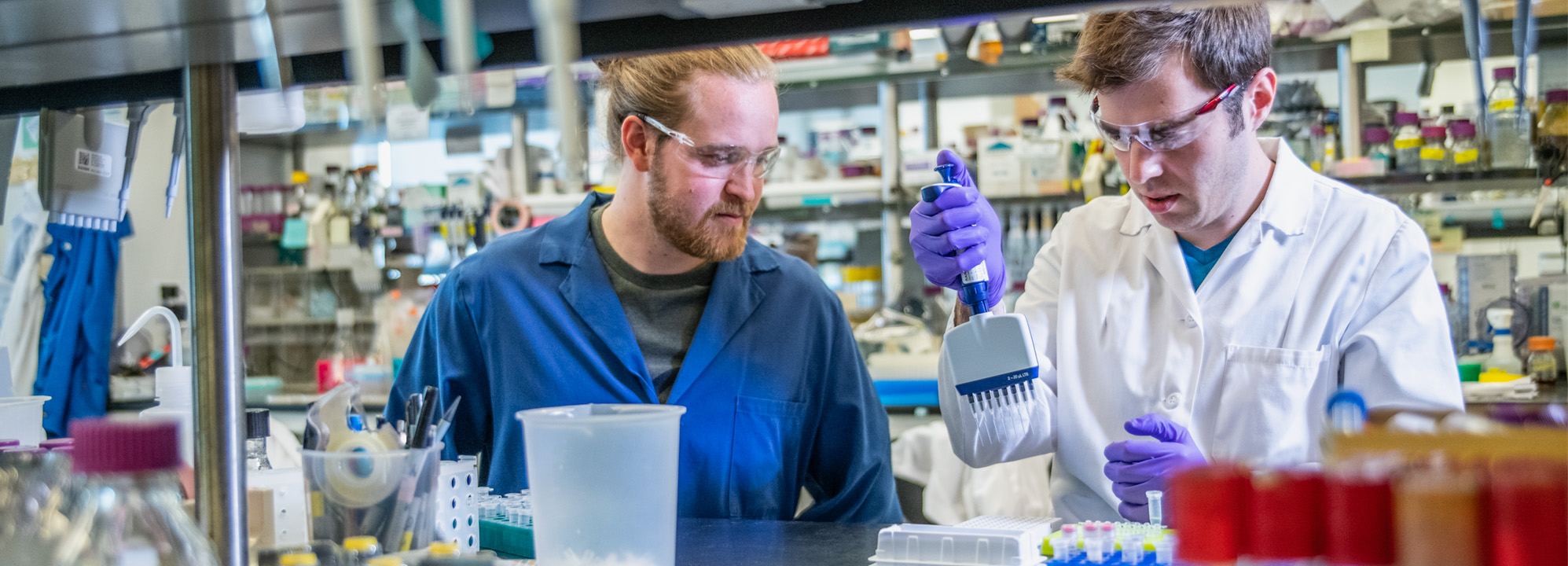 A man in a blue lab coat watches as a colleague in a white lab coat uses a multi-channel pipette in a laboratory. Vials, flasks, and other implements are seen in the foreground, and containers for chemicals fill shelves behind the scientists.