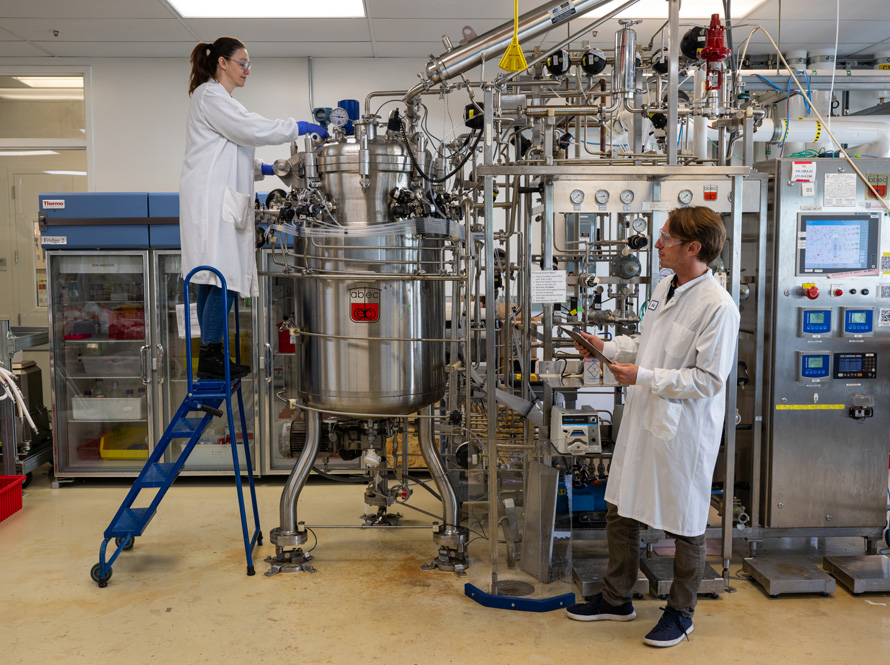 Two researchers wearing white lab coats and safety goggles working with large machinery in the Lab.
