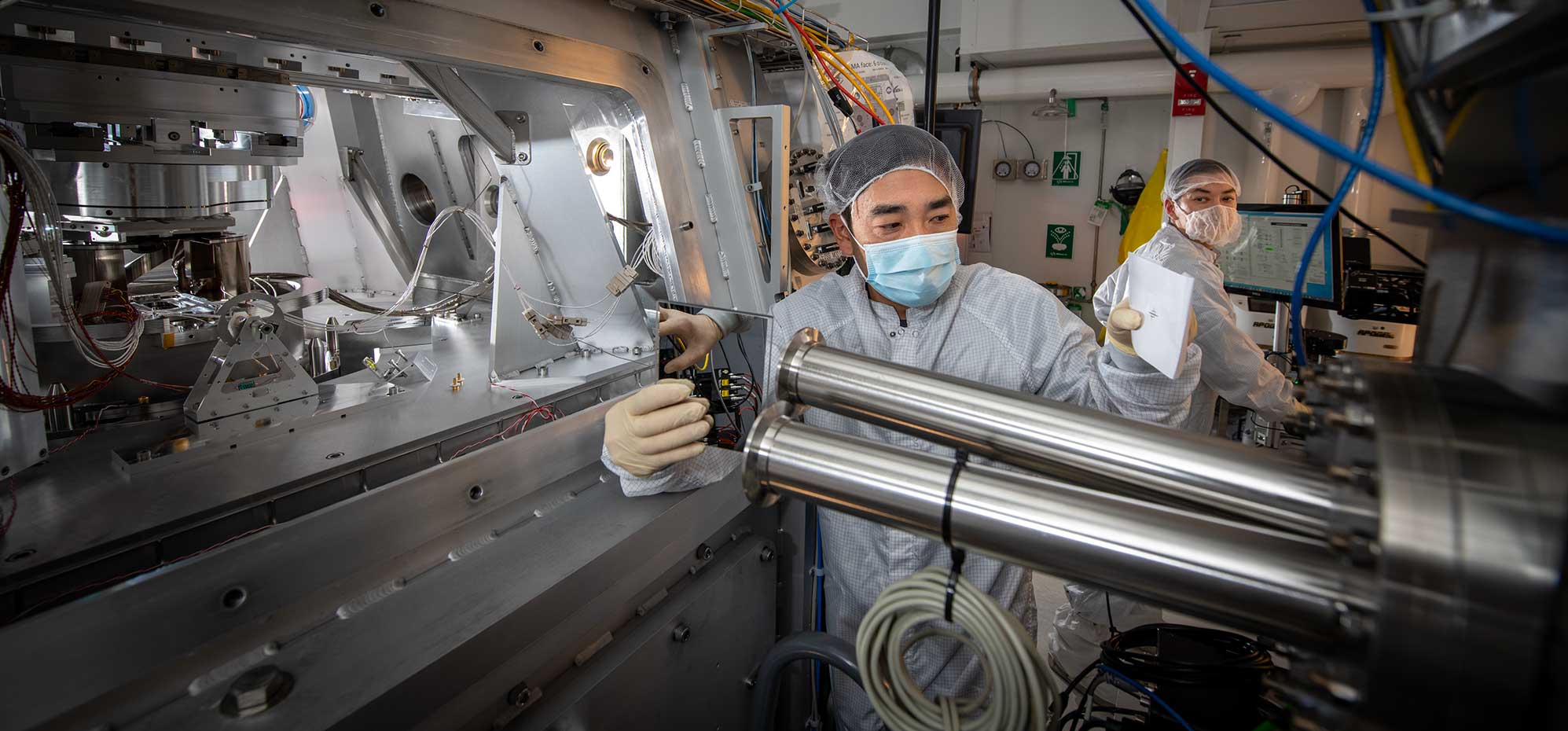 Two researchers wearing white lab coats in a lab with large metal machinery and blue cables.